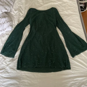 Glittrig grön kortklänning med öppen rygg - Mörkgrön kortklänning med glittriga detaljer, vida långa ärmar och snygg öppen rygg med knytband. Tillverkad i polyester och metalliserade fibrer för en skimrande look. Perfekt för dig som vill sticka ut med en unik och trendig stil.