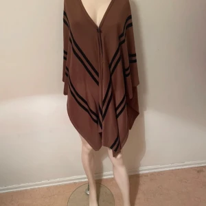 Brun ponchoväst med svarta ränder - Säljer en brun ponchoväst med svarta diagonala ränder och v-ringad hals. Västen har en lös och oversized passform med ett knappfäste framtill. Materialet känns mjukt och lätt, perfekt för lager-på-lager. Snyggt mönster som ger en cool vibe storleken är onesize 