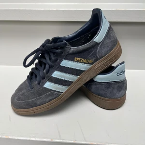 Adidas Spezial sneakers i mörkblå mocka - Snygga Adidas Spezial sneakers i mörkblå mocka med ljusblå detaljer och klassiska tre ränder på sidorna. Skorna har bruna gummisulor, snörning och rund tå. Perfekta för dig som gillar retrostil och streetwear.