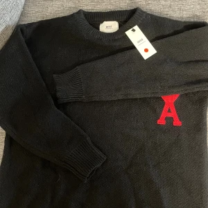 Svart stickad tröja från Ami Paris - Svart stickad tröja från Ami Paris med rund hals och ribbade muddar. På bröstet sitter ett stort rött hjärta med bokstaven A, som är typiskt för märket. Tröjan har en clean och stilren look, perfekt till jeans eller byxor.