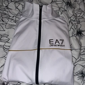 Vit EA7 jacka med svarta detaljer - Snygg vit jacka från EA7 Emporio Armani med svarta dragkedjor och tunna guldiga linjer. Jackan har hög krage, lång ärm och två fickor med dragkedja. Perfekt för dig som vill ha en stilren och sportig look.