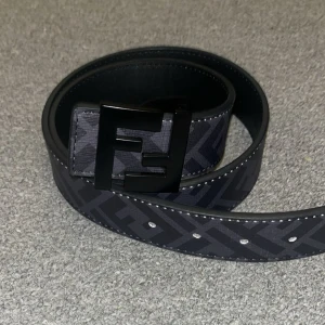Äkta Fendi bälte - Snyggt svart bälte från Fendi med klassiskt FF-mönster och stort svart metallspänne. Bältet är gjort i skinn och har diskreta grå detaljer i mönstret. Perfekt accessoar för att lyfta din outfit med en lyxig touch.
