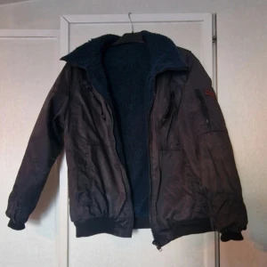 Svart arbetsjacka - Svart arbetsjacka med dragkedja framtill, ribbade muddar och flera fickor med dragkedja. Perfekt för arbete utomhus under kyligare dagar. Välanvänd jacka med skavanker. Tyget är slitet på några ställen och fodret är noppigt och ser slitet ut. Den har använts bland djur så hår förekommer trots att den har tvättats. Jackan har mycket kvar att ge än! Det går inte att se vad det står på storlekslappen men skulle tippa på att det är en stl L.