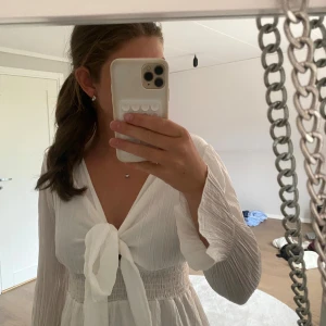 Vit kort omlottklänning med knyt - Superfin vit kort klänning med långa ärmar och knyt framtill. Klänningen har v-ringning, smock i midjan och volangig kjol. Materialet är lätt och luftigt, perfekt för varma dagar. Snygg och trendig modell som ger en romantisk vibe.