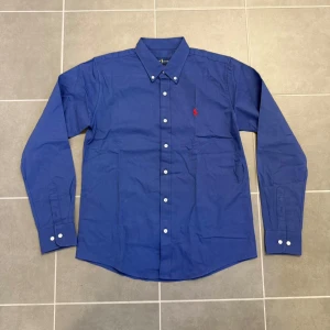 Blå skjorta från Ralph Lauren L - Klassisk blå skjorta från Ralph Lauren i bomull med button-down krage och vita knappar. Broderad röd logga på bröstet. Skjortan har lång ärm och klassisk passform, perfekt för en clean och stilren look.