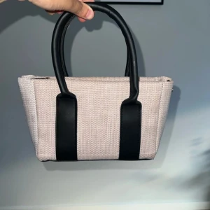 Beige och svart handväska i canvas - Stilren handväska i beige canvas med svarta detaljer och handtag i skinnimitation. Väskan har en rektangulär form, dragkedja upptill och innerfack. Perfekt storlek för det viktigaste och passar till många outfits. Inget band med, om man har det så kan det även användas som en axelremsväska 