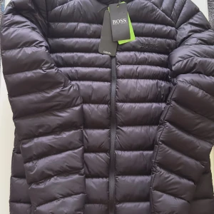 Svart dunjacka från Hugo Boss - Snygg svart dunjacka från Hugo Boss med quiltad design och diskret logga på bröstet samt patch på ärmen. Jackan har dragkedja framtill och är perfekt för kyliga dagar. Klassisk och stilren look som passar till det mesta. Jackan är knappt använd.