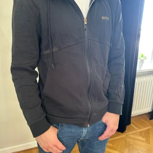 Svart hoodie från Hugo Boss - Svart hoodie från Hugo Boss med dragkedja framtill och guldfärgad logga på bröstet. Tröjan har huva med snörning, ribbade muddar och är tillverkad i mjuk bomull. Snygga detaljer med små svarta nitar vid axelpartiet. Perfekt för en chill och stilren look.