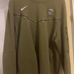 Olivgrön PSG hoodie från Nike - Säljer en olivgrön hoodie från Nike med Paris Saint-Germain-logga på bröstet. Tröjan har dragkedja hela vägen, huva och en extra ficka med dragkedja på ärmen. Perfekt för dig som gillar sportig stil och fotboll.
