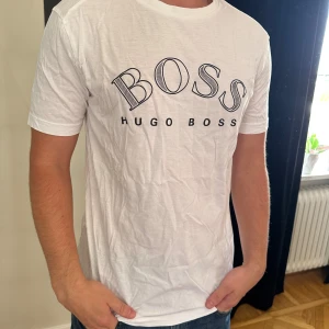 Vit Hugo Boss t-shirt med tryck - Vit t-shirt från Hugo Boss med stort BOSS-tryck i svart på bröstet. Klassisk rund hals och korta ärmar. Tillverkad i mjuk bomull som känns skön mot huden. Perfekt för dig som gillar stilrena och enkla plagg med tydlig logga.