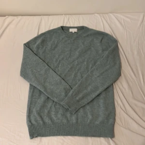 Grå kashmirtröja från Kujten - Supermjuk och stilren grå tröja i 100% kashmir från Kujten. Klassisk rund halsringning, ribbade muddar och lång ärm. Perfekt för dig som vill ha en enkel men lyxig look till höst och vinter.