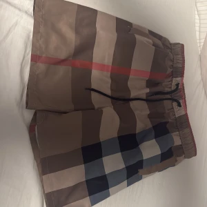 Beige burrberryshorts - Säljer ett par klassiska badshorts från Burberry med ikoniskt rutigt mönster i beige, brunt, blått, svart och rött. Shortsen har elastisk midja med snörning och är tillverkade i ett snabbtorkande material.