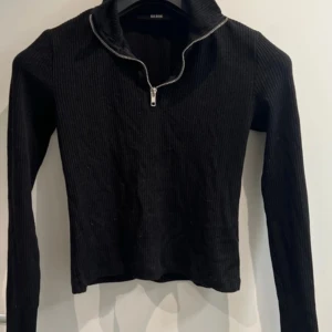 Svart ribbad tröja från BikBok - Svart långärmad tröja från BikBok i ribbat material. Tröjan har en hög krage och en halv dragkedja framtill som ger en cool look. Perfekt att styla med jeans eller kjol för en trendig outfit.