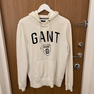 Vit hoodie från Gant med tryck - Snygg vit hoodie från Gant med svart logotyp och sköldtryck framtill. Tröjan har huva med snörning och en stor magficka. Perfekt för dig som gillar klassisk och sportig stil.