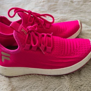 Rosa sneakers från Fila - Snygga och sportiga sneakers från Fila i en stark rosa färg. Skorna har stickad ovandel i syntetmaterial, snörning och vit platt sula. Perfekta för dig som vill sticka ut med färg och gillar bekväma skor till vardags eller träning.