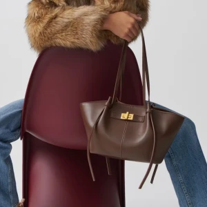 Brun axelväska i skinn med gulddetalj - Stilren brun axelväska i skinn med långa remmar och snygg guldfärgad vridknäppning framtill. Väskan har en mjuk, något strukturerad form och minimalistisk design med diskreta sömmar och dekorativa knytband på sidorna. Perfekt för dig som gillar klassiska accessoarer.