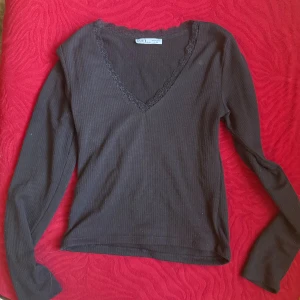 Svart ribbad långärmad topp Karol basic - Svart ribbad långärmad topp från Karol basic med v-ringning och spetskant runt halsen. Storlek L men känns som en M.Toppen har en slim passform och är tillverkad i ett mjukt, stretchigt material som sitter snyggt på kroppen. Perfekt att styla med jeans eller kjol för en clean look.