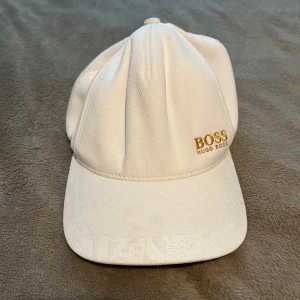 Vit keps från Hugo Boss - Snygg vit keps från Hugo Boss med broderad logga i guld framtill. Justerbar rem baktill med metallspänne där det också står BOSS. Kepsen är i klassisk modell och tillverkad i ett strukturerat bomullstyg. Perfekt accessoar för en clean och stilren look.