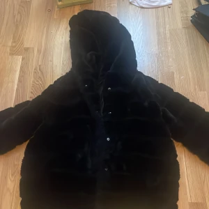 Svart pälsjacka med huva - Mysig svart pälsjacka med huva och tryckknappar framtill. Jackan har lång ärm och är fluffig med en riktigt skön känsla. Perfekt för kalla dagar när du vill vara både varm och snygg.