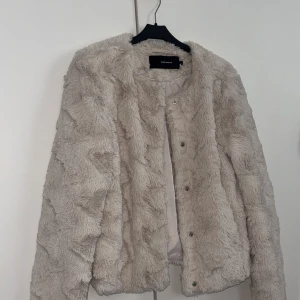 Beige pälsjacka från Vero Moda - Supermjuk och fluffig beige pälsjacka från Vero Moda. Jackan har rund halsringning, tryckknappar framtill och är helfodrad. Perfekt för dig som vill ha en mysig och trendig look under kyliga dagar.
