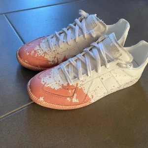 Maison Margiela sneakers med färgstänk - Unika sneakers från Maison Margiela i vitt med rosa färgstänk över tå och sidor. Modellen har rund tå, platt sula och klassisk snörning. Tillverkade i canvas med coolt konstnärligt mönster som sticker ut. Perfekta för dig som gillar statement pieces och kreativ stil.