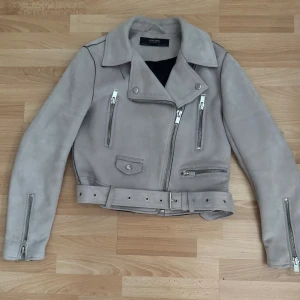 Grå imitation mocka bikerjacka från Zara - Snygg grå bikerjacka från Zara Basic i mjuk imitation mocka med coola silverfärgade dragkedjor, flera fickor och bälte i midjan. Jackan har klassisk krage och långa ärmar med dragkedjor vid ärmslut. Perfekt för dig som gillar en edgy stil.