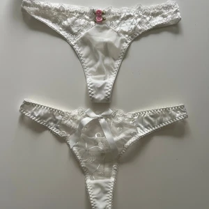 Vit spetsbikini med rosett och blommor - Säljer en vit bikini med spetsdetaljer och transparent mesh. Överdelen har två små rosa blommor framtill och underdelen har en söt vit rosett och broderade blommor. Båda delarna har smala band och en feminin, elegant look.