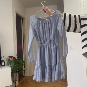 Ljusblå prickig klänning med volang - Supersöt ljusblå klänning med små vita prickar, långärmad och med volang nertill. Klänningen har resår i midjan för en snygg passform och är gjord i ett lätt, tunt material som känns luftigt och skönt.