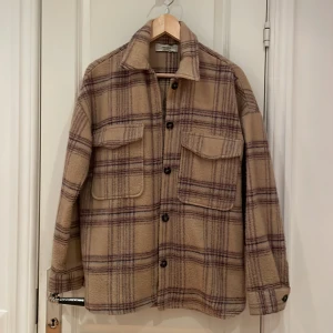 Beige rutig overshirt från Pull&Bear - Säljer en beige och lila rutig overshirt från Pull&Bear. Jackan har två stora bröstfickor med lock, knäppning framtill och mjukt, ulligt material. Perfekt för lager-på-lager och har en avslappnad passform med långa ärmar.