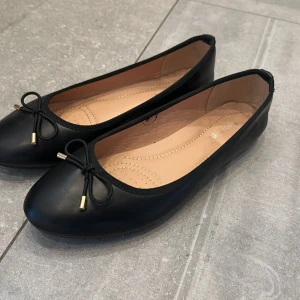 Svarta ballerinaskor från Andarina - Klassiska svarta ballerinaskor från Andarina med rund tå och dekorativ rosett framtill med gulddetalj. Skorna har en platt sula och är tillverkade i syntetmaterial med mjuk innersula för extra komfort. Står storlek 41 men de är små i storlek så skulle säga mellan 39-40.