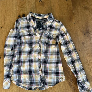 Rutig blus från Guess Jeans M - Snygg figursydd blus från Guess Jeans, M/38. Mkt sparsamt använd. Tryckknappar, tre knappar på ärmslutet. Möjlighet att kavla upp ärmen och fästa. Snörning i ryggen,  liten ficka på framsidan. Vit botten, ljusgula och svarta streck och rutor, tunna röda linjer. Supersnygg!