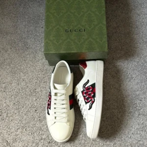 Gucci Ace sneakers med orm - Gucci Ace sneakers i vitt skinn med klassiska gröna och röda ränder på sidan och broderad röd orm. Baksidan har metallicröd och metallicgrön detalj med Gucci-logga. Snygg och ikonisk modell som sticker ut.