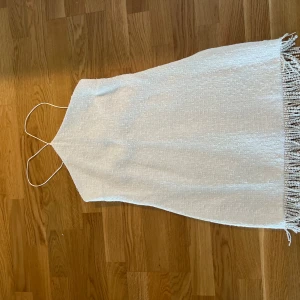 Vit kort klänning med pärlfrans - Supersöt vit kort klänning med tunna axelband och pärlfrans längs nederkanten. Klänningen har en lätt skimrande struktur och är fodrad upptill med slät satin. Perfekt för dig som vill sticka ut med en unik detalj och stilren look.