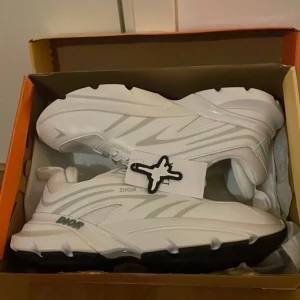 Vita  sneakers chunky modell - Säljer ett par helt nya vita chunky sneakers med svart sula och snygga detaljer. Skorna har snörning, meshpaneler och tydlig -logga på plös, häl och sidor. Perfekta för dig som gillar statement sneakers med modern vibe.