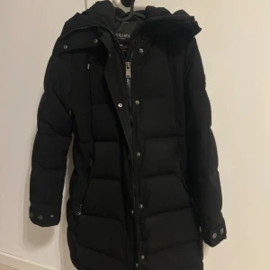 Svart lång pufferjacka Hollies 34 - Svart lång pufferjacka från Hollies med huva, dragkedja och knappar framtill. Finns även en avtagbar päls till jackan som jag har tagit av, men om de önskas kan man få med den. Skriv privat för mer bilder. Använd 1 vinter så den är i bra skick