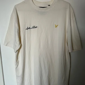 Vit t-shirt från Lyle & Scott Archive - Vit t-shirt från Lyle & Scott Archive med boxy fit. T-shirten har korta ärmar, rund hals och broderad logga i gult på bröstet samt svart text. Tillverkad i mjuk bomull, perfekt för en chill och clean look.