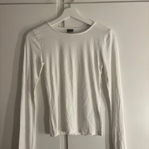 Vit långärmad topp från Gina Tricot - En basic vit långärmad topp från Gina Tricot i mjuk bomull. Enkel och stilren design med rund halsringning och figurnära passform. Perfekt att styla med jeans eller kjol för en clean look.