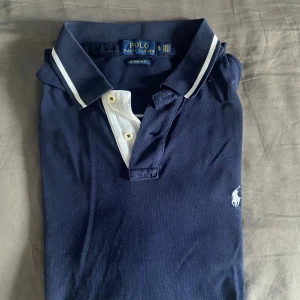 Marinblå Polo Ralph Lauren piké XL - Marinblå pikétröja från Polo Ralph Lauren i bomull, custom slim fit. Klassisk krage med vita ränder, knappar framtill och broderad logga på bröstet. Stor vit siffra 2 på ryggen och vänster ärm.