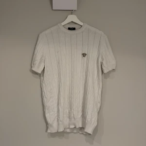 Vit kabelstickad tröja från Versace - Vit kabelstickad tröja från Versace med korta ärmar och rund hals. Tröjan har ett broderat Versace-emblem på bröstet och ribbade muddar vid ärmslut och nederkant. Perfekt för dig som vill ha en lyxig och stilren look.