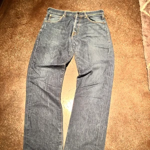 Evisu jeans med broderade bakfickor - Unika blå jeans från Evisu med ikoniska broderade bakfickor i rött, vitt och blått. På fickorna står det 'WE ARE WITH EVISU' i röd text. Klassisk passform, raka ben och snygga kontrastsömmar. Jeans i bomull med läderpatch i midjan och cool streetkänsla.