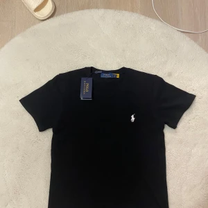 Svart t-shirt från Polo Ralph Lauren - Svart t-shirt från Polo Ralph Lauren med klassisk vit broderad logga på bröstet. T-shirten har rund halsringning och korta ärmar. Tillverkad i mjuk bomull för en skön känsla och snygg passform.