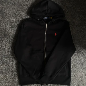Svart hoodie från Polo Ralph Lauren - Svart hoodie från Polo Ralph Lauren med dragkedja och huva. Klassisk röd logga broderad på bröstet. Tröjan har långa ärmar, ribbade muddar och känguruficka framtill. Perfekt för en chill och stilren look.