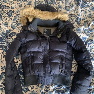 Mörkblå pufferjacka med fuskpäls - Snygg mörkblå pufferjacka från American Eagle med huva som har vit fordring och fuskpäls. Liknar de eftersökta jackorna från abercrombie & fitch och från Hollister!! Inga defekter som jag kan hitta!! Passar perfekt till hösten. Jag bär storlek XS som preferens och jag skulle säga att den är storlek XS-M men det står M på taggen. Skriv för fler bilder!