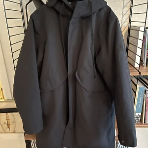 Svart vattentät parka från Tretorn XL - Svart parka från Tretorn i storlek XL. Jackan har huva med snörning, stora fickor framtill och dragkedja med vindslå. Tillverkad i återvunnen nylon från fiskenät, en del av Sea Life Trust-kollektionen. Perfekt för kalla och blöta dagar.