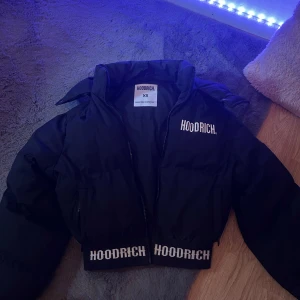 Svart pufferjacka från Hoodrich XS - Svart pufferjacka från Hoodrich i storlek XS. Jackan har en croppad passform, dragkedja framtill och tydliga vita Hoodrich-loggor på bröstet och längs nederkanten. Perfekt för dig som vill ha en streetstyle-look med attityd.