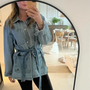 Jeansjacka med bälte och stora fickor - Snygg jeansjacka i ljusblå denim med knappar framtill, stora bröstfickor och midjebälte för en trendig siluett. Jackan har klassisk krage och långa ärmar, perfekt för lager-på-lager-stil.