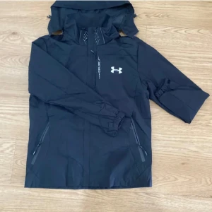 Svart vindjacka från Under Armour - Svart vindjacka från Under Armour med vit logga på bröstet och diskreta detaljer vid dragkedjan. Jackan har huva, meshfoder och två fickor med dragkedja. Perfekt för blåsiga dagar och sportiga outfits. Står L men känns mer som M