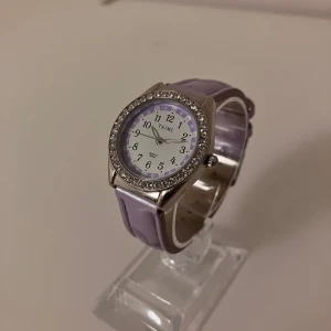 Lila klocka med blommigt armband från TAJMS - Snygg klocka från TAJMS med lila armband i syntetmaterial och blommigt mönster. Urtavlan är vit med lila detaljer och silverfärgade siffror. Runt boetten sitter glittrande stenar som ger extra bling. Perfekt accessoar för dig som gillar färg och detaljer. Behöver ett nytt batteri .