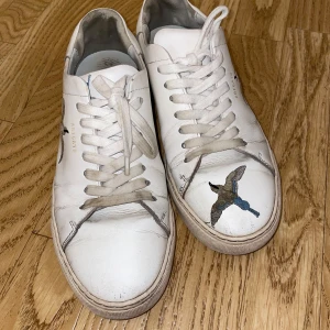 Vit sneaker från Axel Arigato med broderi - Snygga vita sneakers från Axel Arigato med broderade fåglar i blått och beige på sidorna och tån. Skorna har klassisk rund tå, platt sula och snörning. Tillverkade i skinn med diskret guldtext på sidan. Mycket använda
