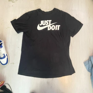 Svart Nike t-shirt Just Do It L - Svart t-shirt från Nike i storlek L med klassiskt 'Just Do It'-tryck i vitt på bröstet. Skön passform och tillverkad i mjuk bomull. Perfekt för dig som gillar sportiga och stilrena plagg med ikonisk logga.
